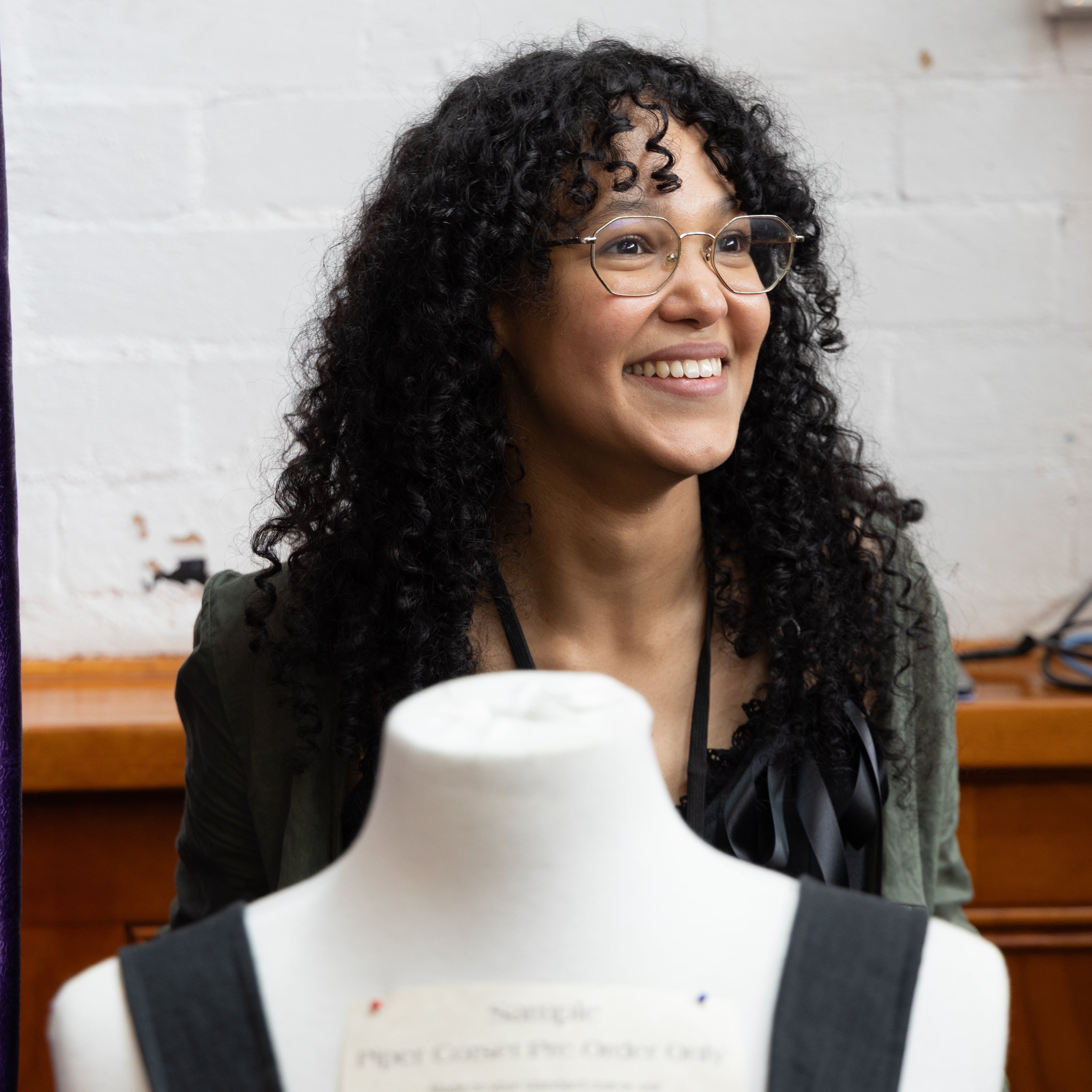 Woman standing next to a mannequin wearing a black garment in a room with purple fabric and wooden furniture.